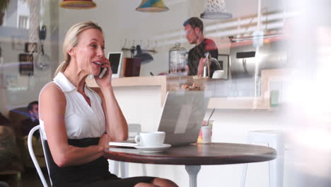 Mujer-De-Negocios-En-Una-Mesa-En-Un-Café-Usando-Un-Teléfono-Inteligente-Y-Una-Computadora-Portátil