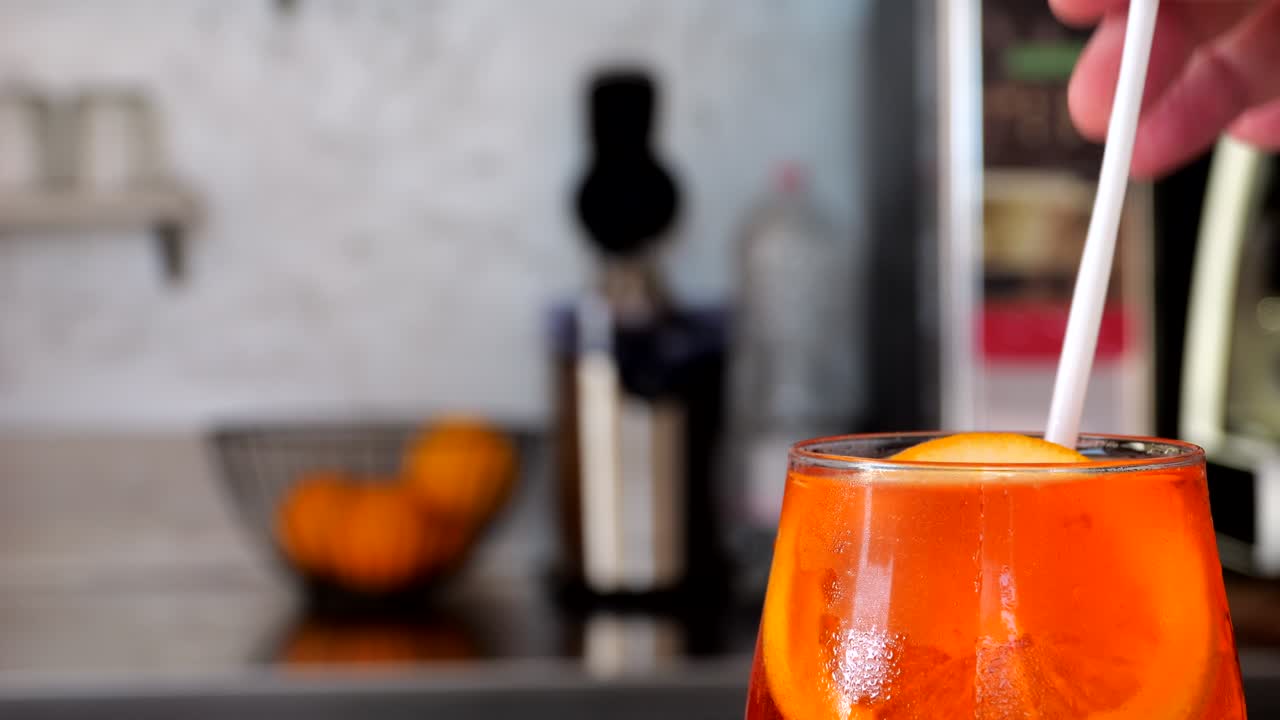 Hand stirs a bright orange Spritz Veneziano in a stemmed glass, garnished with an orange slice, against a blurred bar counter background