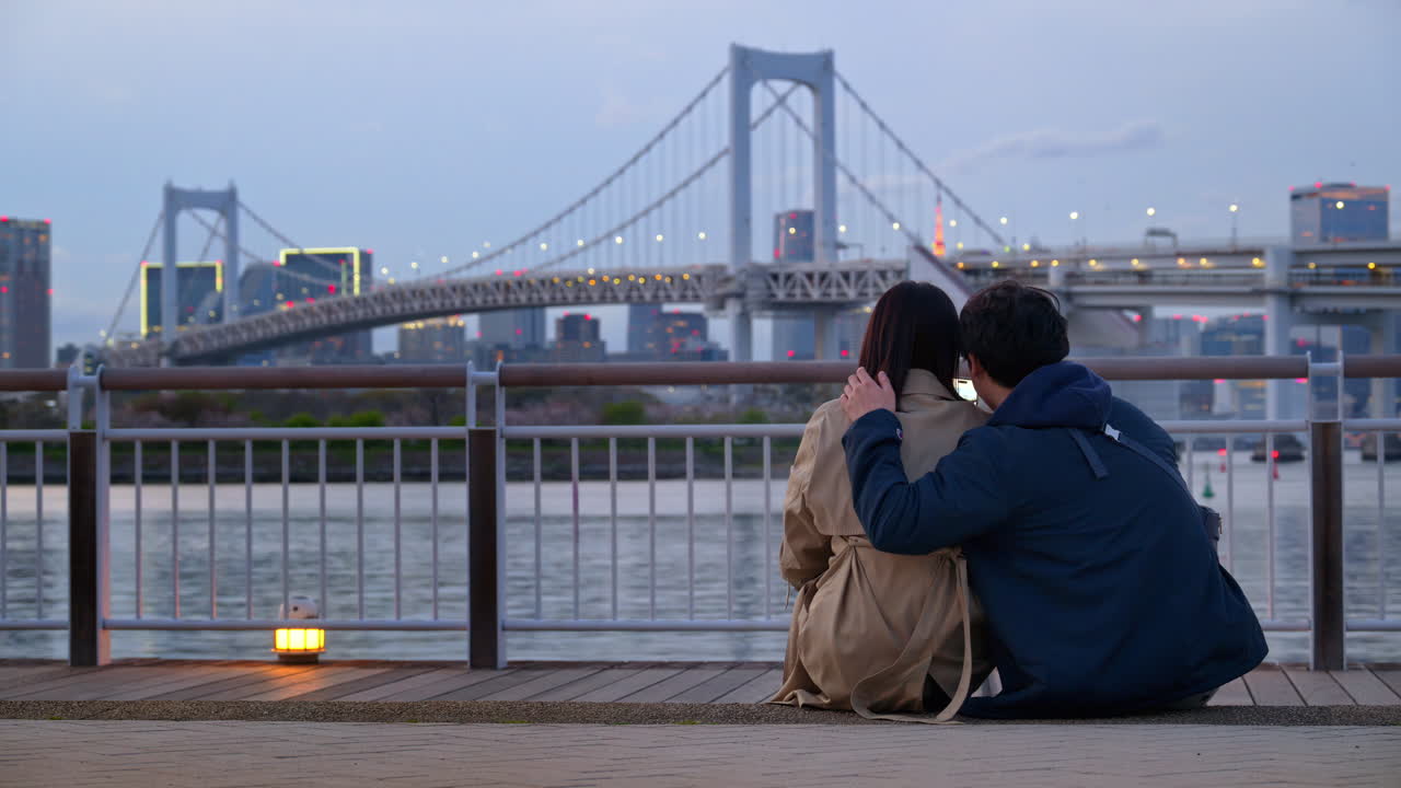 A man and a woman couple sitting in front of a view of the Rainbow Bridge and the skyline of Tokyo, Japan in the evening