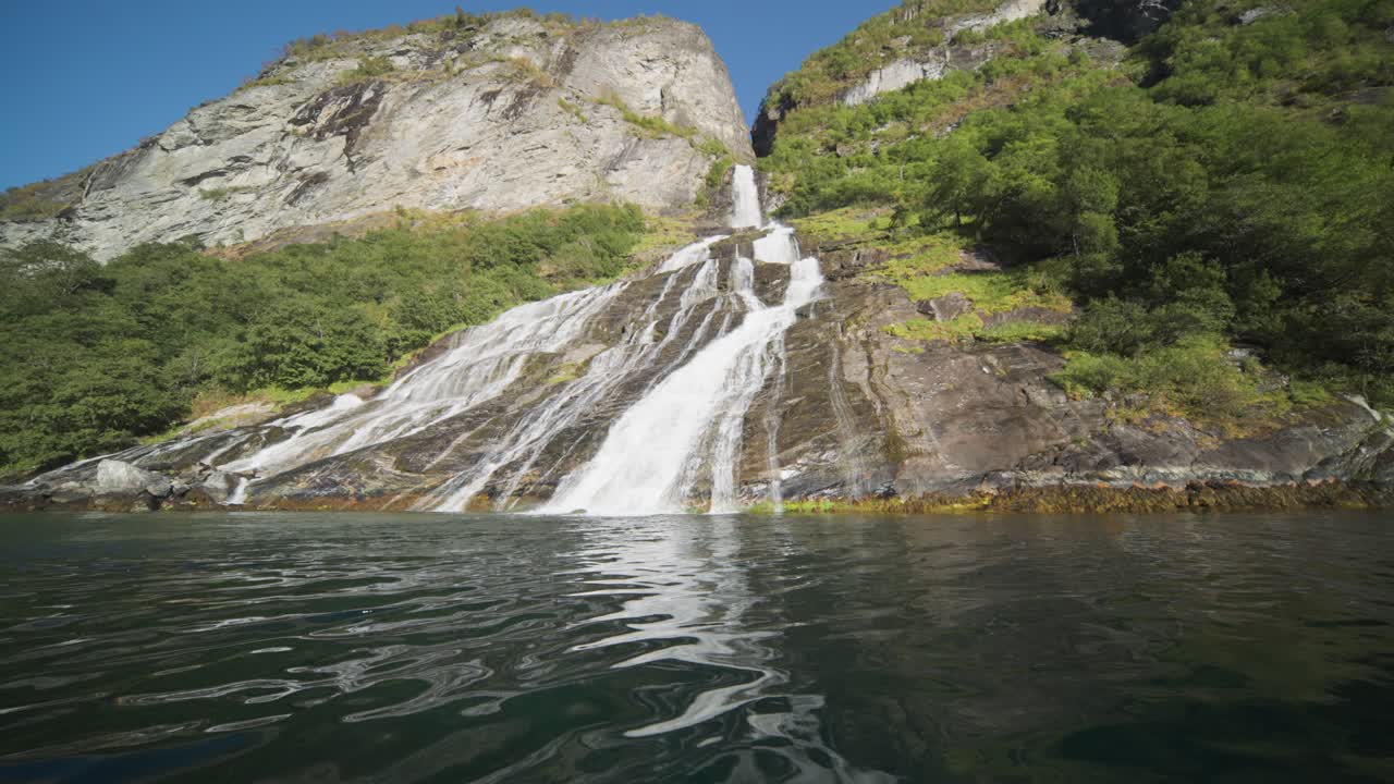A close-up view of a cascading waterfall flowing into the calm waters of Geiranger Fjord, surrounded by lush greenery on the rocky shores. An over-under view.