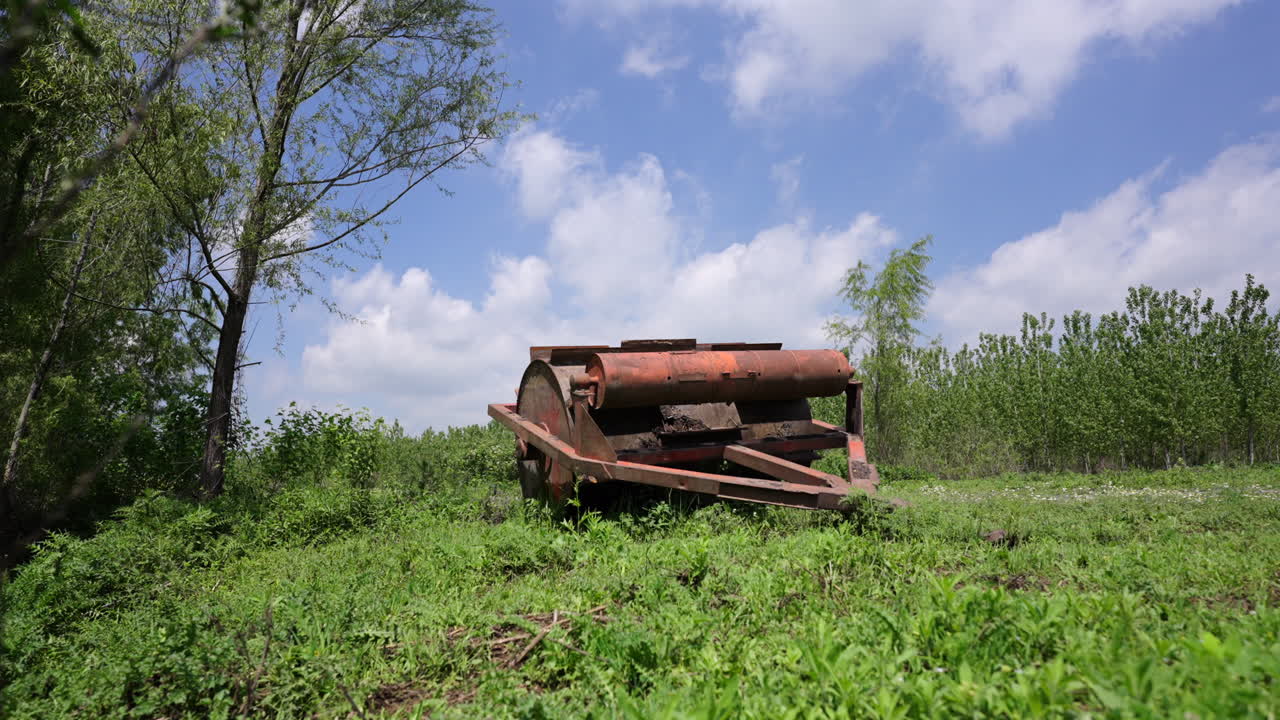 Old rusty road roller abandoned in a field with green grass and trees under a blue sky with white clouds, representing decline, decay and abandonment