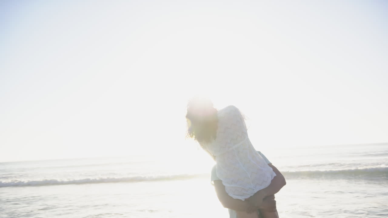 이중 인종 부부는 복사 공간과 함께 빛이 빛나는 해변에서 장난스러운 순간을 즐기고 있습니다.