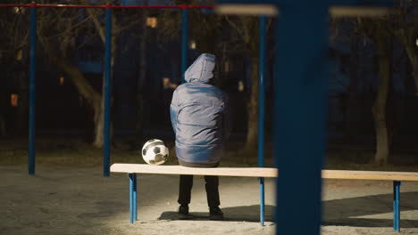 vista trasera de una persona con una chaqueta con capucha azul sentada en un banco al aire libre por la noche con una pelota de fútbol colocada a su lado, con reflejo de luz de los alrededores