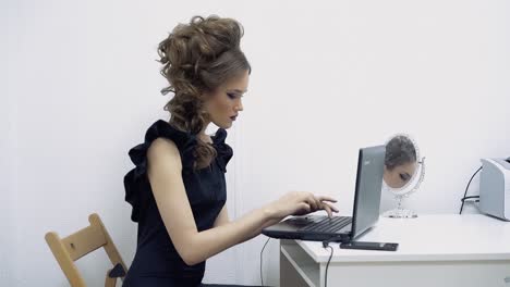 Young-girl-in-the-office-office-manager-sitting-at-the-laptop-typing-text-sometimes-looking-at-phone