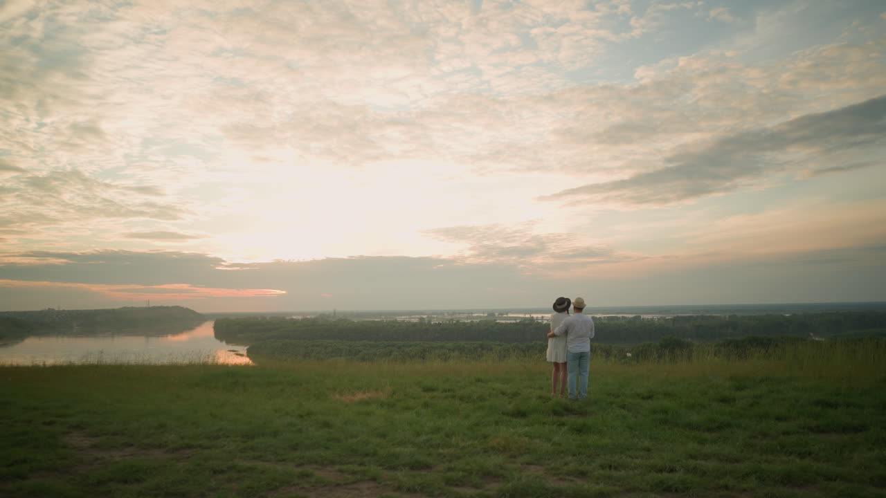 한 부부는 해가 지는 동안 호수 의 잔디 언덕 위에 서 있습니다. 남자는 색 셔츠, 모자, 청바지를 입고 여성의 허리를 잡고 있습니다. 그녀는 검은 모자와 색 드레스를 입고 있습니다.