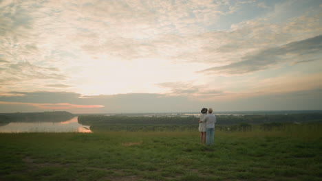 a couple stands on a grassy hill beside a lake during sunset, viewed from behind. the man, wearing a white shirt, hat, and jeans, holds the woman by the waist. she wears a black hat and a white dress