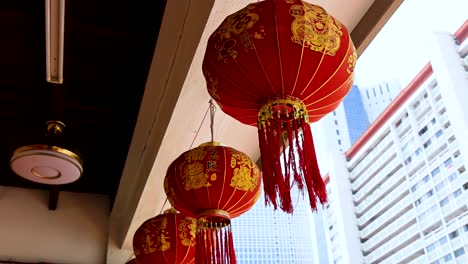 red chinese lanterns hang and gently sway under a covered walkway, with tall modern buildings in the background, captured in bright natural daylight