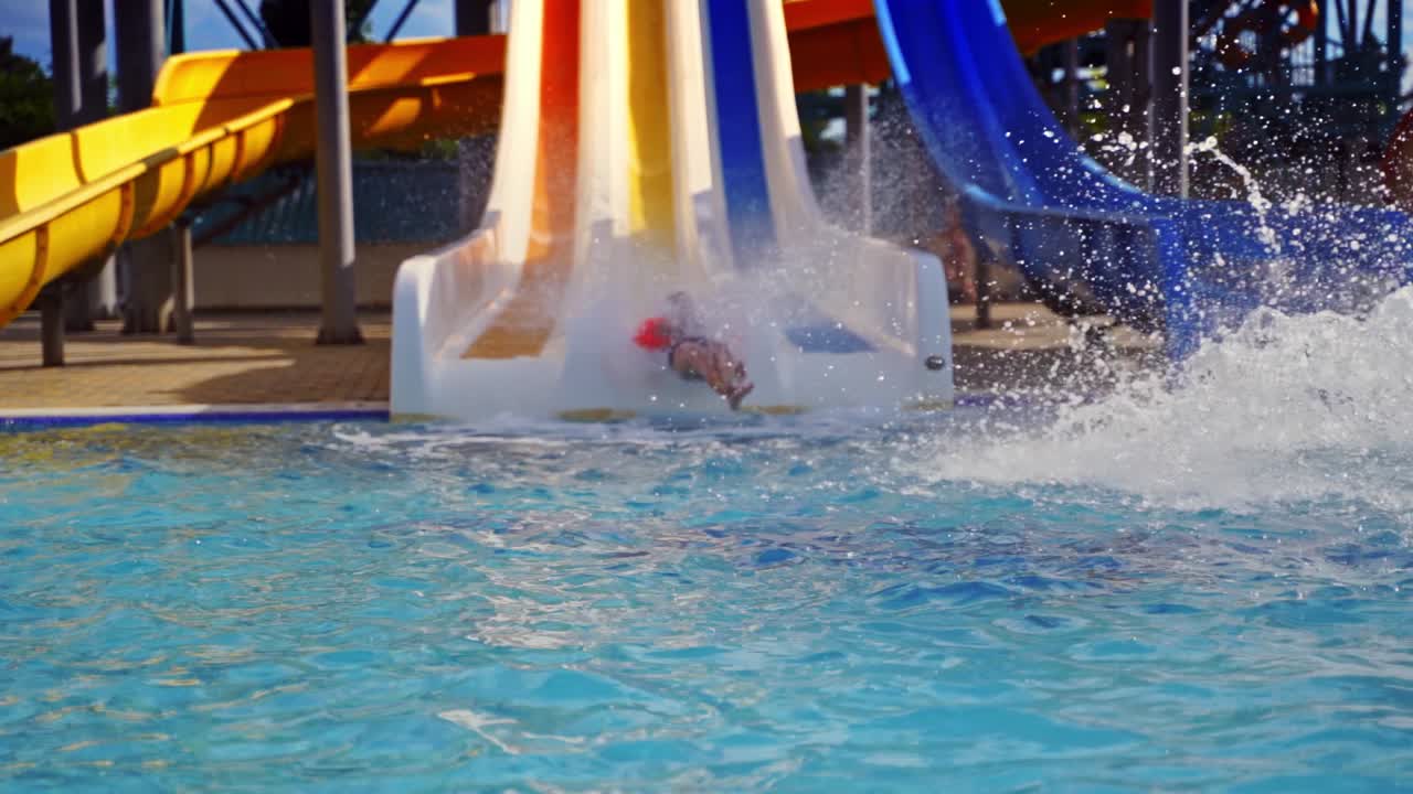 Colorful outdoor water park in summer. Cute boy riding on a slider in the water park. Water splash in the pool. Fun and happiness. Slow motion.