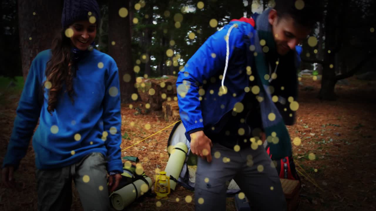Una coppia in viaggio arriva in una radura nel bosco con gli zaini, sistemando l'attrezzatura accanto a una tenda gialla per il campeggio.
