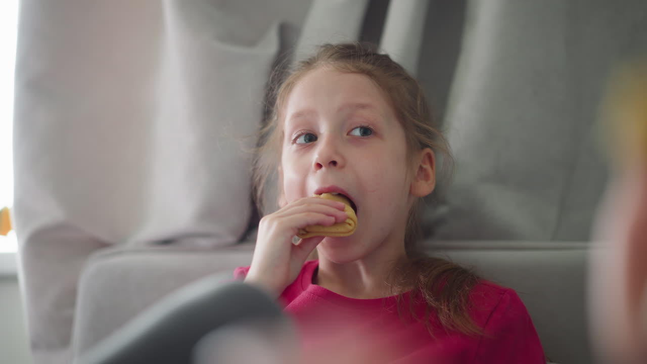 Little girl wearing red top sitting indoors savoring snack while using hand to push food inside mouth, looking directly at camera with relaxed expression, blur view of another person in background