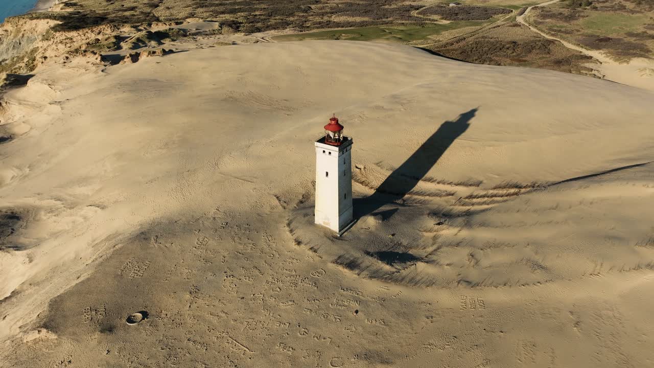 A stunning drone shot capturing Denmark's iconic stone lighthouse standing tall beside large sand dunes on the coast, showcasing the raw beauty and contrast of nature and historic architecture.
