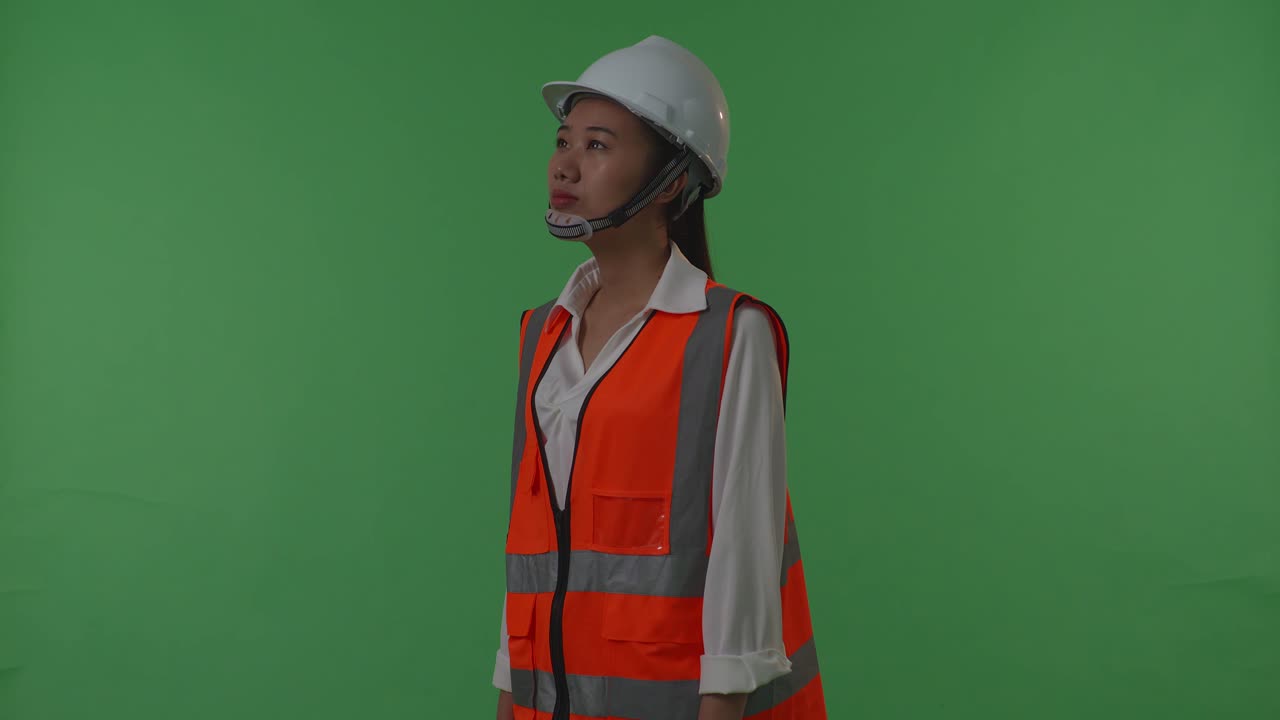 vista lateral de una ingeniera asiática con casco de seguridad mirando a su alrededor mientras está de pie en el estudio de fondo de pantalla verde, durante la puesta o el amanecer