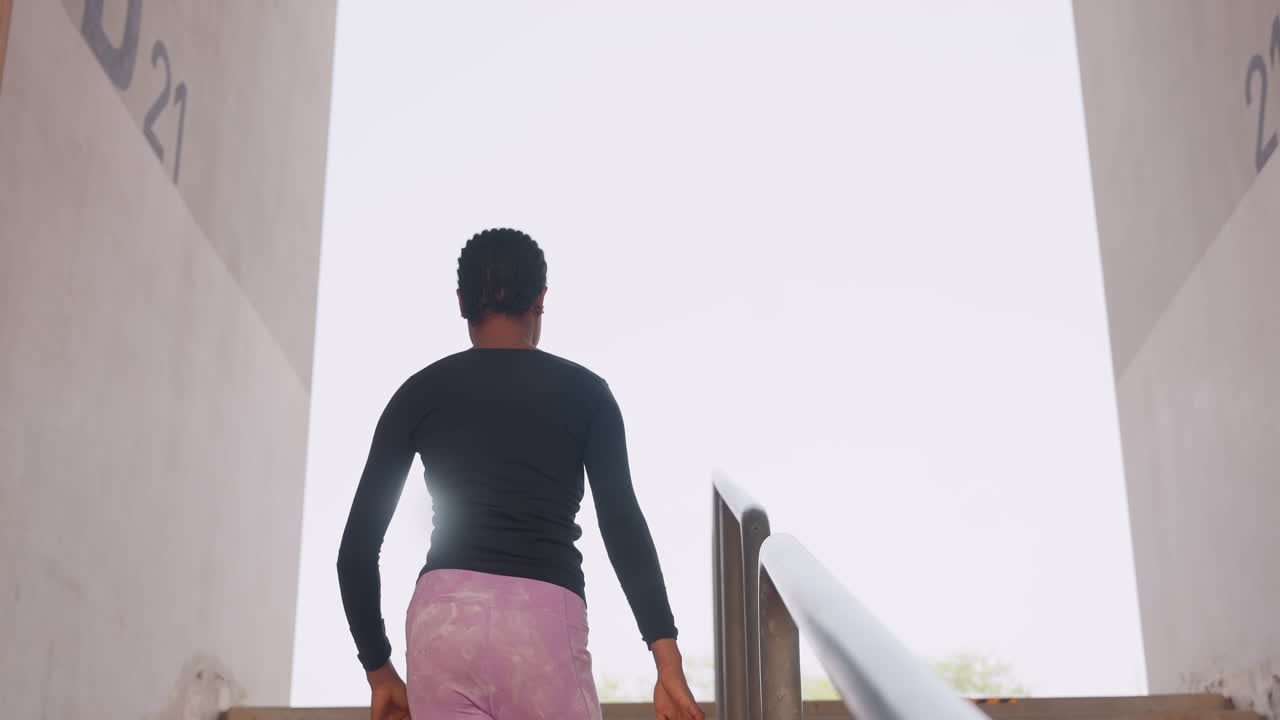 black woman walking into bright opening at top of stairs from rear, silhouette against light, steady ascent conveying transition and quiet optimism, minimal architecture and airy horizon framing new