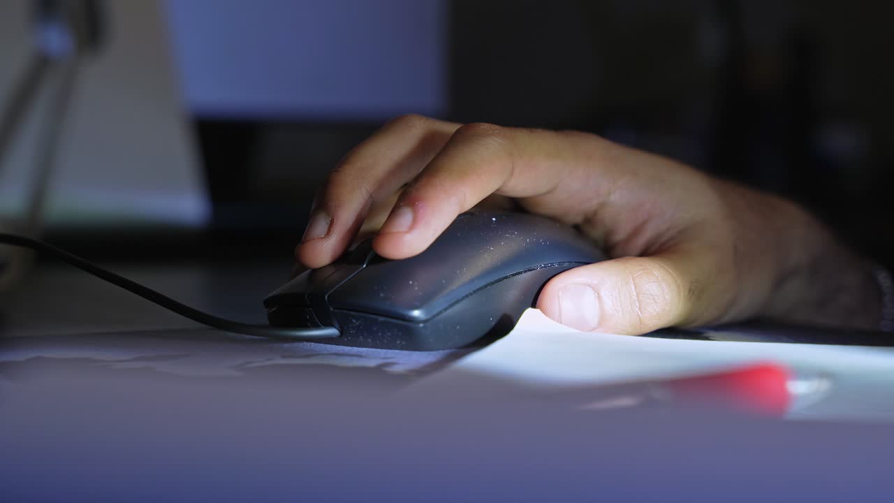 a hand using a wired computer mouse on a desk in low light, with shallow focus, paper details, and an indoor workspace atmosphere