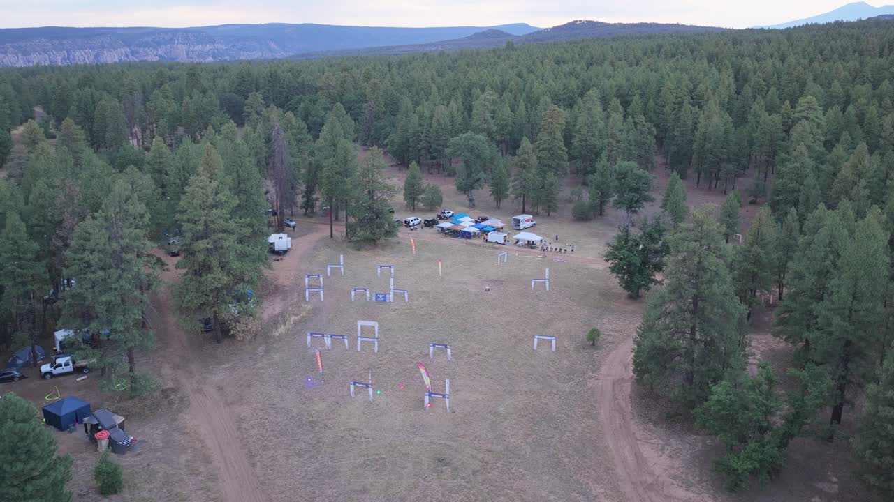 Ilmakuva metsän raivaamisesta iltahämärässä drone-kilparadan ollessa valmiina. Teltat, ajoneuvot ja varusteet näkyvissä. Tapahtuman valmistelukohtaus hämärtyvässä päivänvalossa. Moottoriurheilupaikan yläkuva