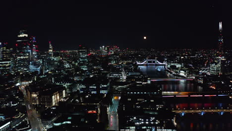 Backwards-fly-above-historic-city-centre-at-night.-Tall-modern-skyscrapers-in-background.-London,-UK