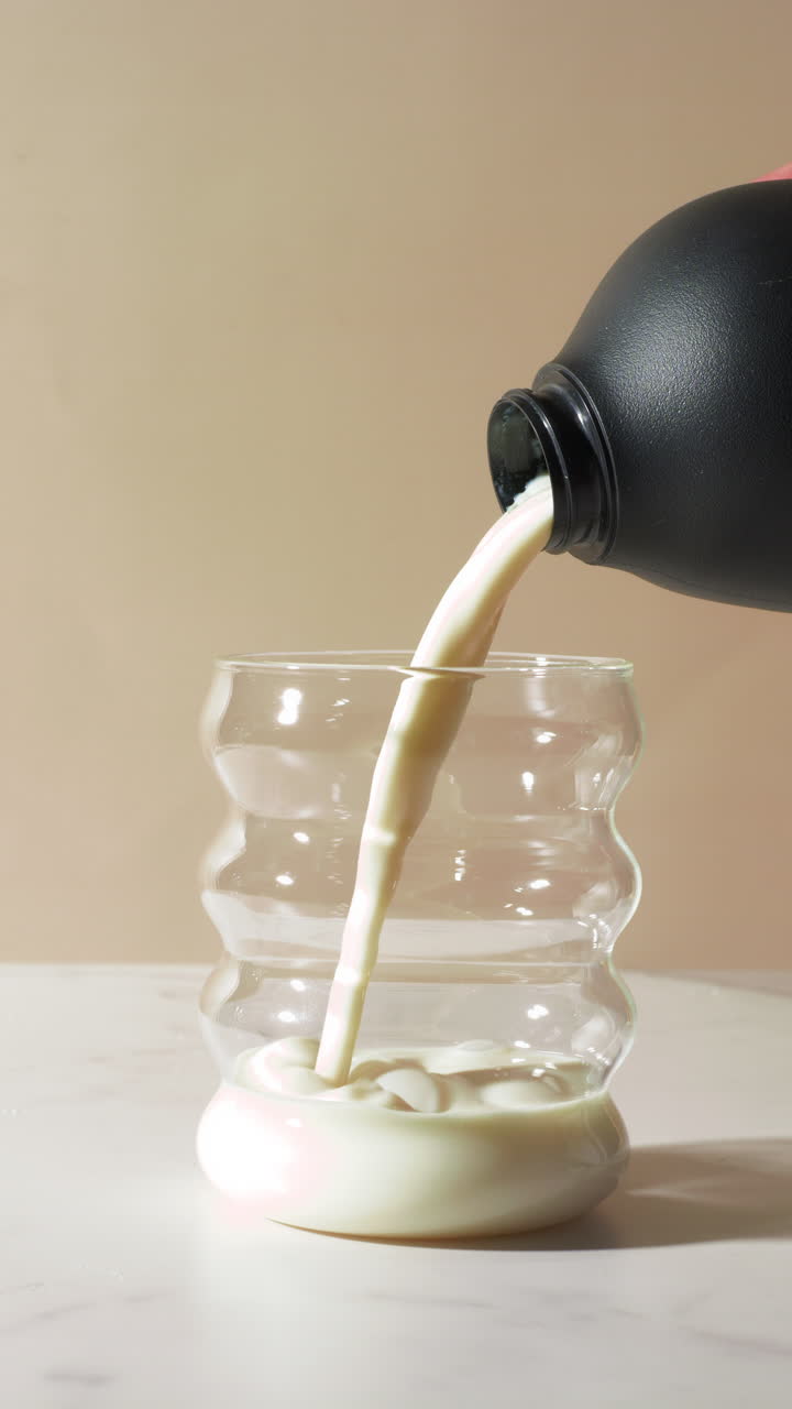 Slow-motion close-up of a thick vanilla or chocolate protein shake being poured from a black bottle into a clear, wavy glass. Creamy texture, smooth flow emphasize nutrition, energy, meal replacement.