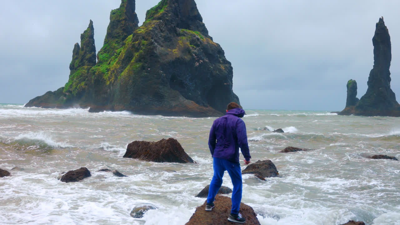 hombre con chaqueta azul de pie sobre una roca manteniendo el equilibrio en la playa de arena negra de reynisfjara en islandia en un día ventoso
