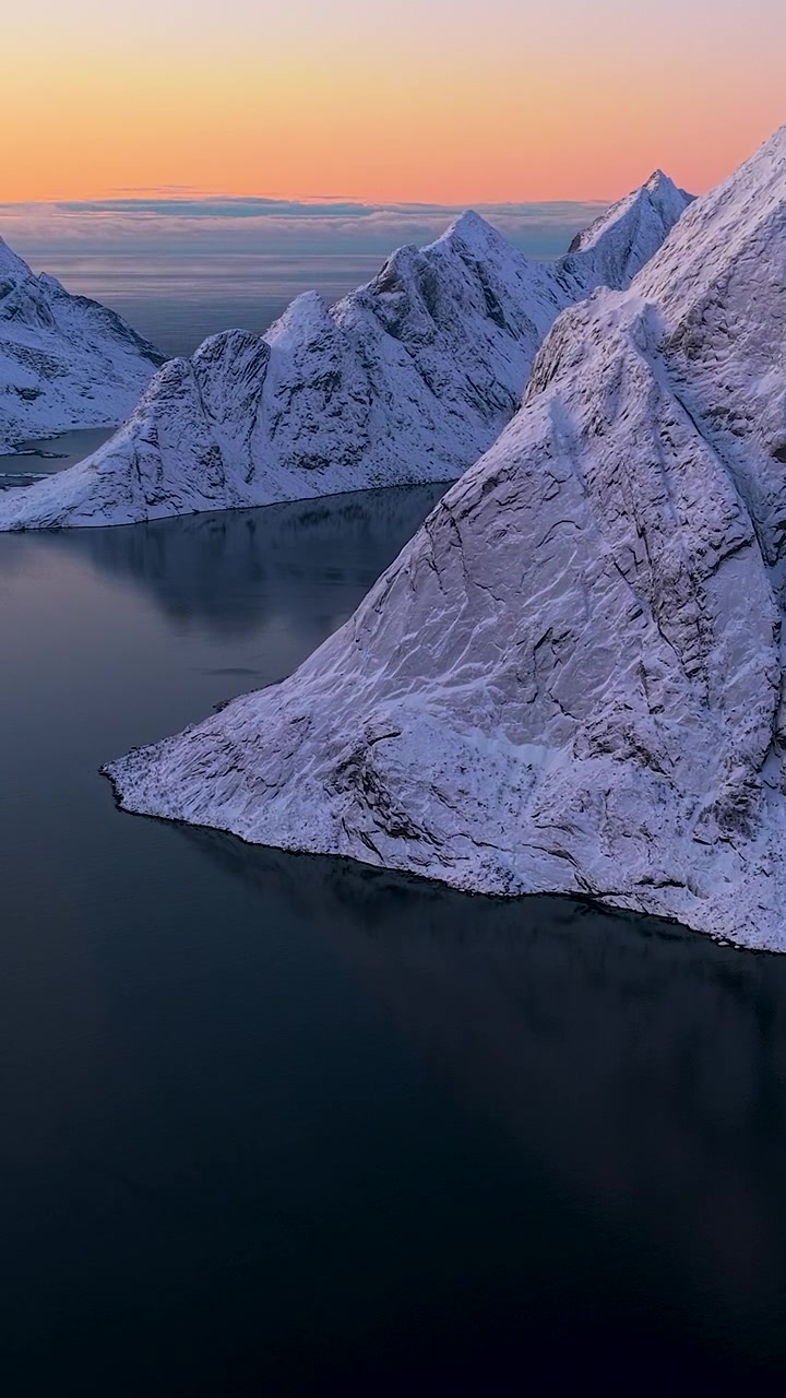 Atemberaubende vertikale Drohnenaufnahme von schneebedeckten Berggipfeln, die auf das dunkelblaue Fjordwasser auf den Lofoten, Norwegen, während eines lebhaften Sonnenuntergangs treffen.