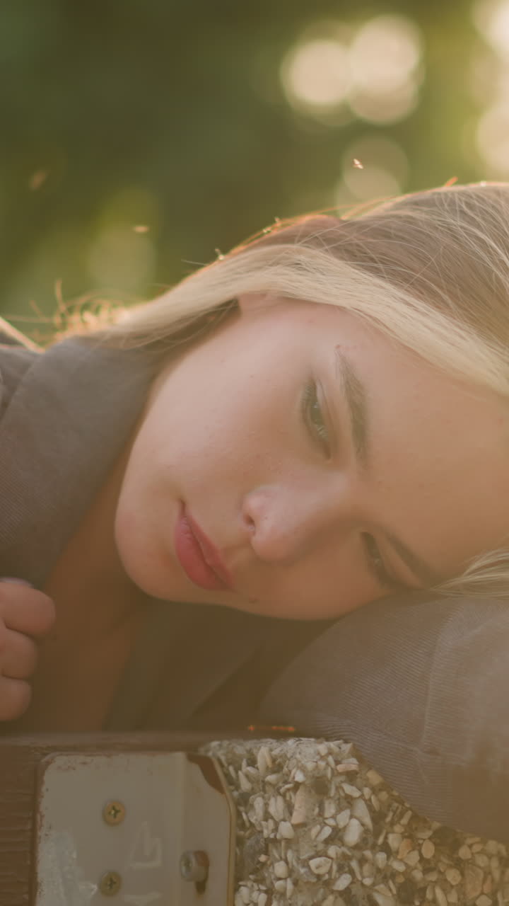 Woman resting her head on arm while seated on bench, gently touching her hair as she drops the hand, soft sunlight creates a warm, peaceful ambience around her, with blurred background