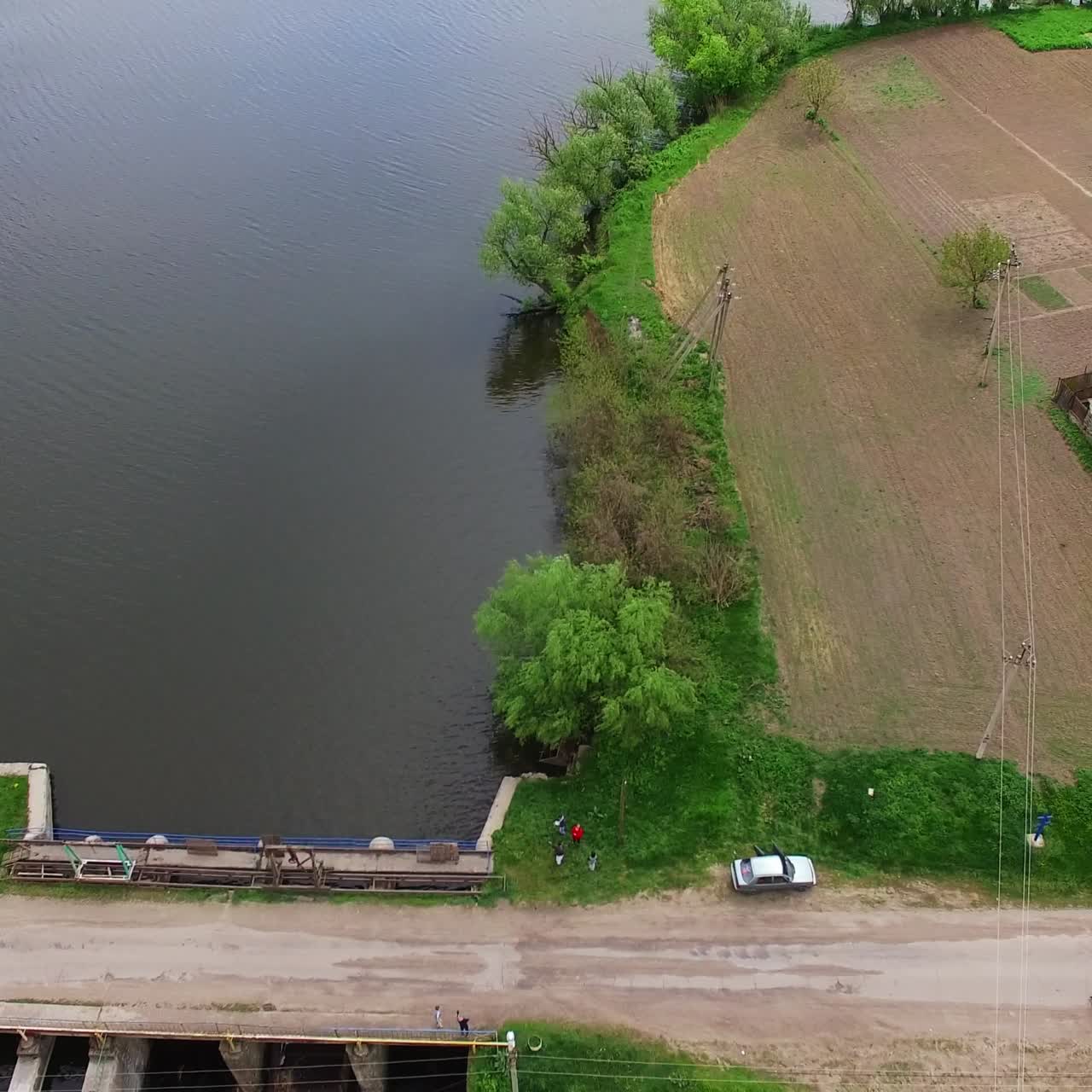 Quick flight over the narrow river with green banks, dam over it. Beautiful farmlands at backdrop. Aerial perspective