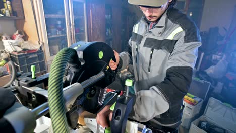 a young man with a beard in gray overalls by profession a carpenter works with a circular cutting machine in his home workshop