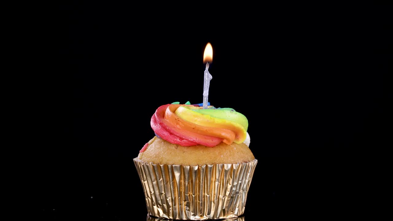 Twelve rainbow-frosted cupcakes with lit candles arranged in a grid pattern, flames gently flickering against a seamless black background under controlled studio lighting