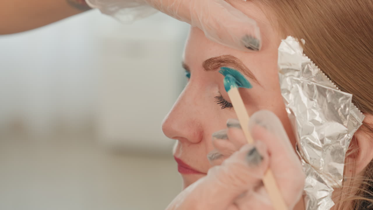 eyebrow tinting teal dye female client, esthetician applies pigment with small brush, foil protects hairline, gloved hands, closeup profile shot, soft neutral lighting, careful precision strokes