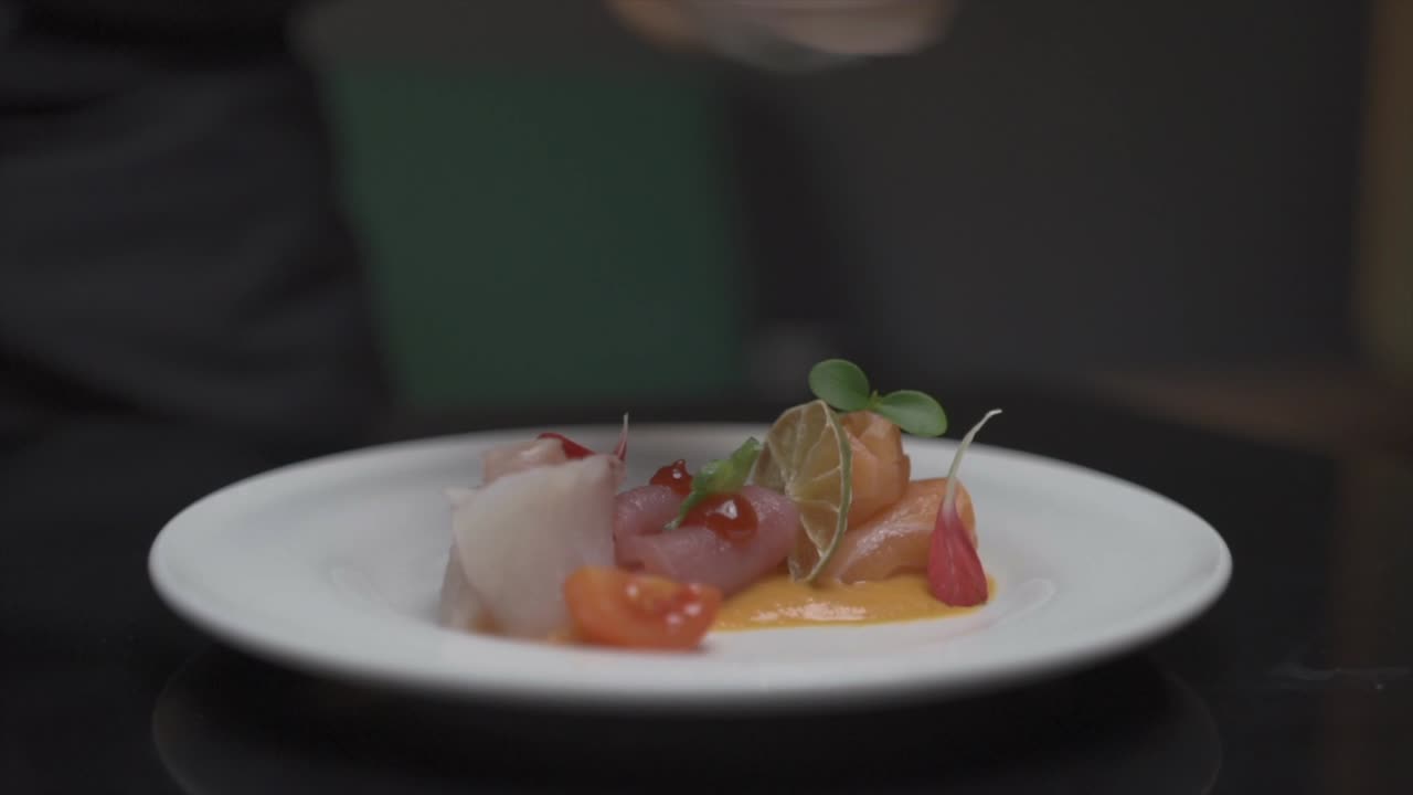 Nærbilde av en autentisk rett hvor kokken pynter en nyberedt japansk sashimirett ved hjelp av grønne urter og blomsterblader, med en krydret dipp.