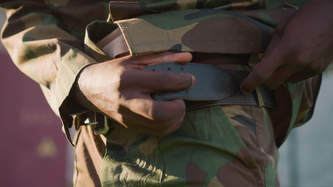 Black Soldier Fastening Belt And Adjusting Uniform With Hands Closeup, Camo Pants And Gear Pouches Visible, Careful Preparation And Tactile Focus Before Movement, Intimate Detail Shot