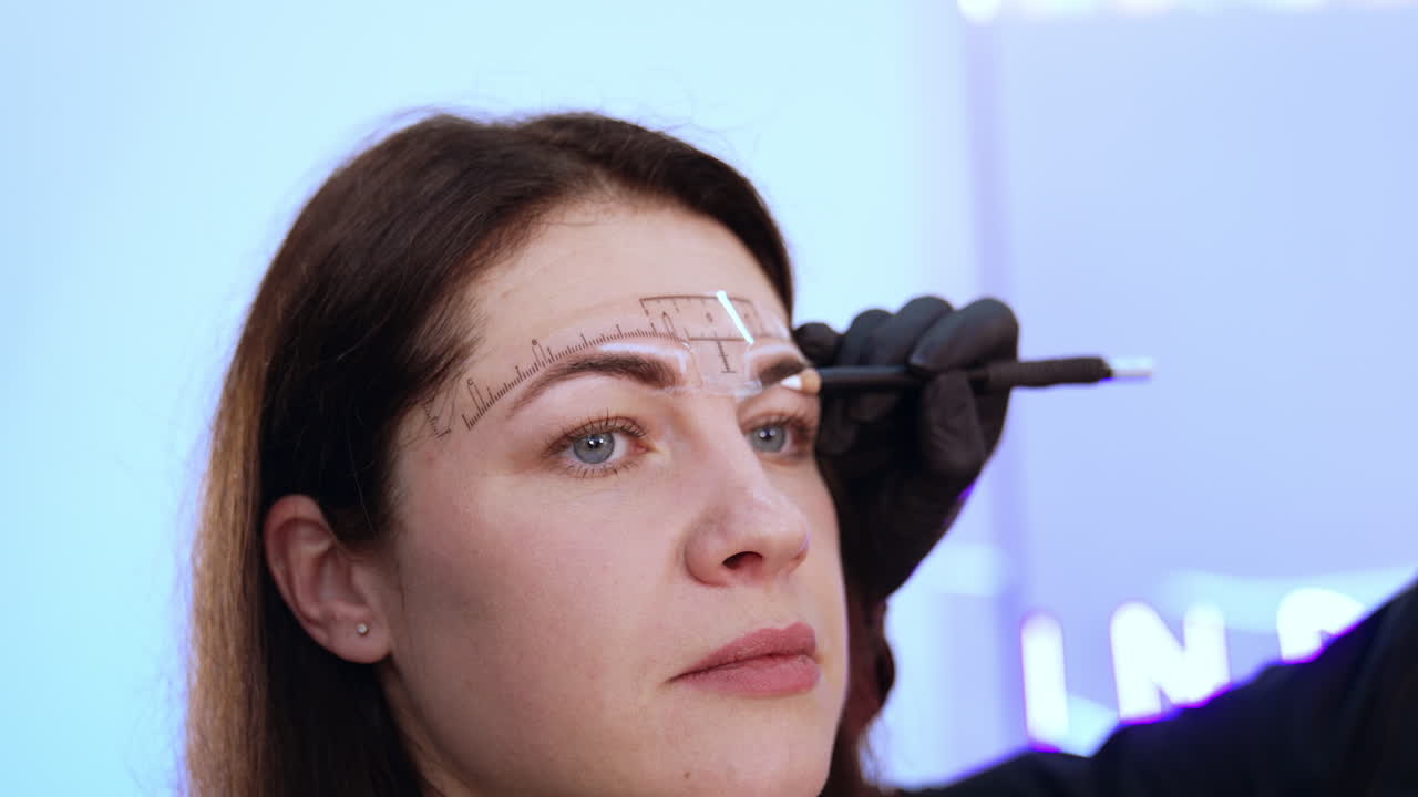 Beautiful adult brunette is having the eyebrow correction procedure. Cosmetologist drawing white lines to mark the limits of eyebrow make up.