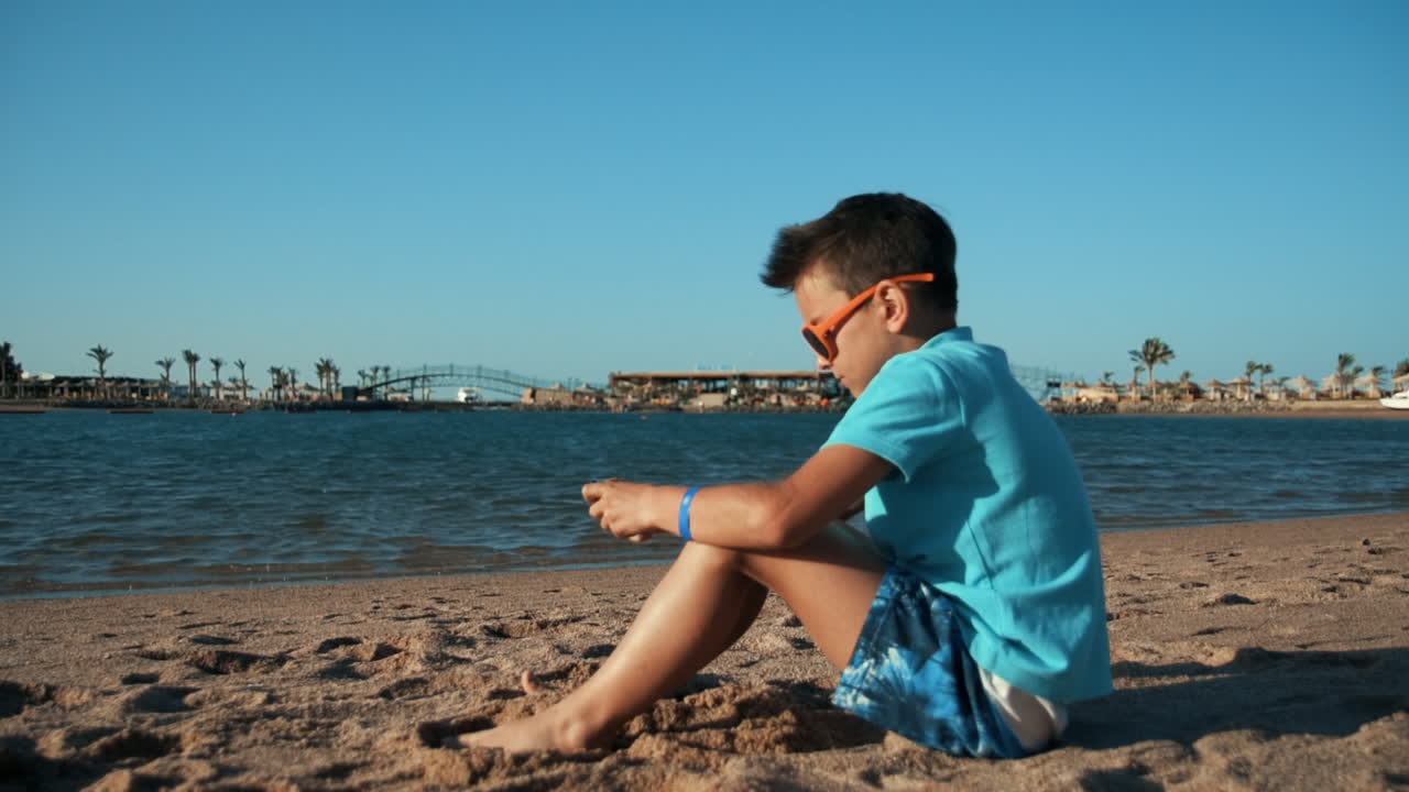 선글라스를 입은 예쁜 젊은 남자가 모래 해변에서  ⁇ 드폰으로 놀고 있습니다.