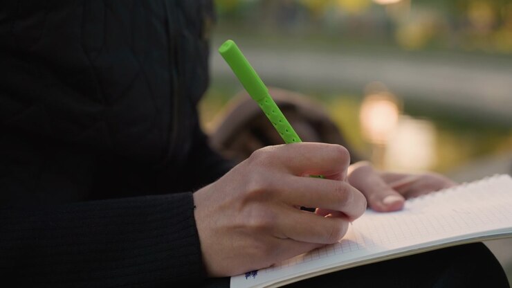 Close up shows person hands holding bright green pen and actively filling pages of open lined notebook. background is softly blurred suggesting outdoor or natural light with warm tones that