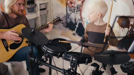 Ein-Glücklicher-Albino-Junge-Mit-Weißem-Haar-In-Einem-Braunen-Pullover-Spielt-Auf-Einem-Elektronischen-Schlagzeug-Mit-Seinem-Blonden-Vater-Mit-Brille-Und-Bart,-Der-An-Seinem-Freien-Tag-Mit-Seinem-Kleinen-Sohn-Im-Zimmer-Akustikgitarre-Spielt