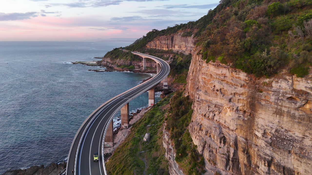Breathtaking aerial shot of a vehicle moving along the Sea Cliff Bridge, surrounded by dramatic cliffs and the blue Pacific Ocean. Revealing aerial shot.