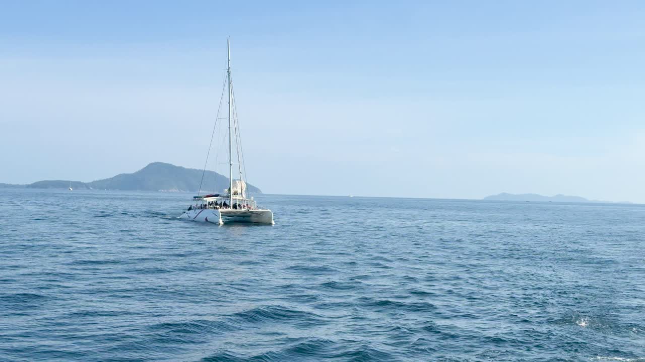 A catamaran sails calmly across Chalong Bay, Phuket, under clear skies with distant islands visible. The scene captures serene oceanic beauty