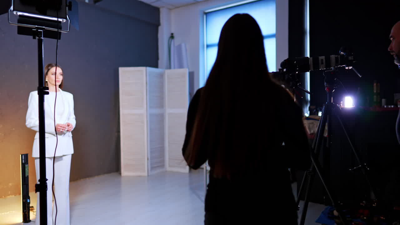 Production crew silhouettes watching female presenter in white suit. Behind the scenes view of studio set with woman in white suit and camera operators in shadow