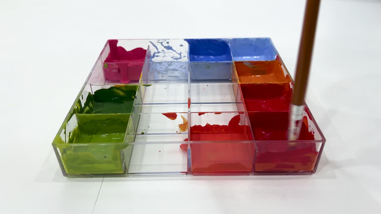 A paintbrush stirs vibrant poster paints in a transparent tray under bright, even lighting. Camera remains steady, focusing on color mixing process