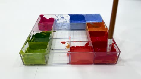 a paintbrush stirs vibrant poster paints in a transparent tray under bright, even lighting. camera remains steady, focusing on color mixing process