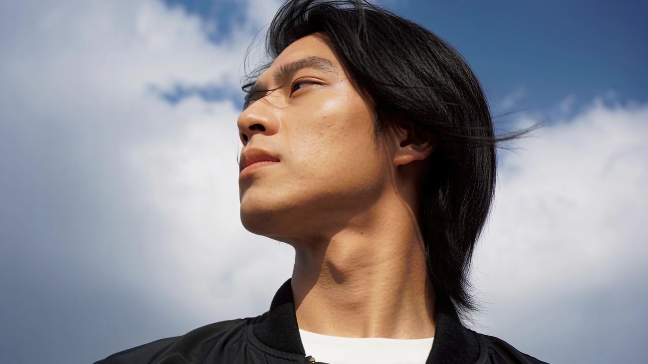 空の下、顎を上げ、顔を向けた男性。風になびく髪。黒のボンバージャケットと白いTシャツを着用。