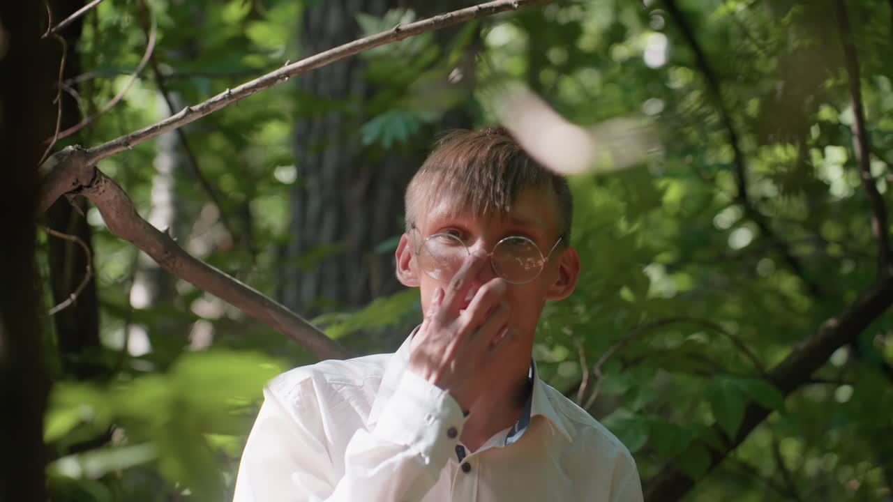 Portrait view of young ecologist in white shirt and glasses standing in forest observing trees with thoughtful expression, sunlight filtering through dense foliage