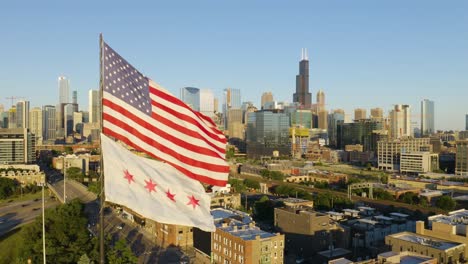 Vista-Aérea-Fija-De-La-Bandera-Estadounidense-Y-La-Bandera-De-Chicago-Ondeando-En-Un-Día-Ventoso-Con-Vista-Del-Horizonte-De-Chicago-En-Segundo-Plano