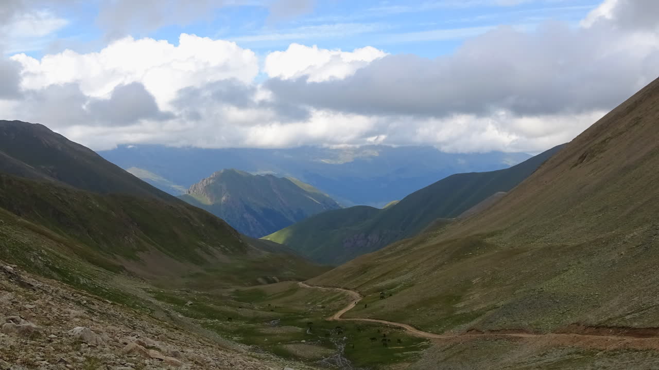 Filmato in timelapse di un'ampia valle di montagna con una strada sterrata tortuosa e nuvole drammatiche sopra pendii verdi, girato nelle montagne del Caucaso, Russia.