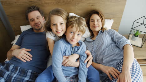 draufsicht auf ein süßes mädchen, das ihren jüngeren bruder umarmt, während sie zwischen ihren glücklichen eltern auf dem bett sitzen