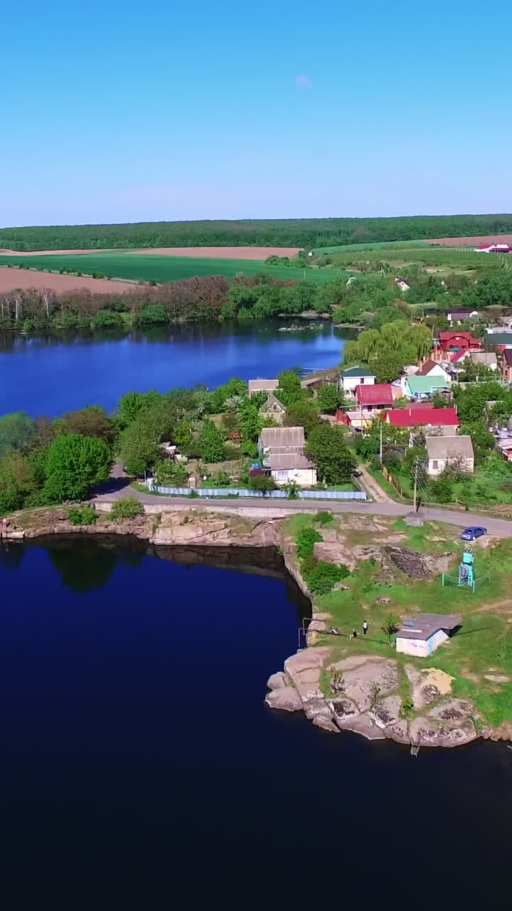 Contrasting azure surface of the river near the green bank. Cute cozy village with beautiful houses on bright sunny daytime from top. Vertical video