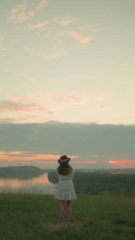 vista trasera de una esposa en un vestido blanco y sombrero de pie en un campo tranquilo de hierba, mirando a un lago tranquilo bajo un cielo suave y de color pastel durante la puesta de sol, rodeado de calma y serenidad