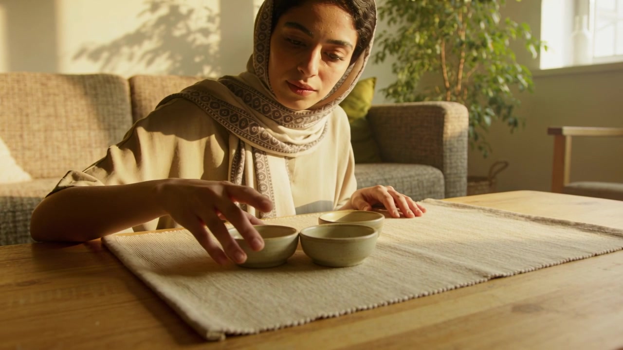 Leaning woman wearing printed hijab beige top stacking three bowls at living room table