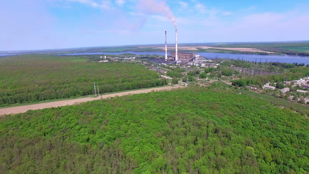 Flying over abundant green forest closer to an industrial zone. Narrow river and agricultural fields at backdrop.