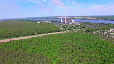 flying over abundant green forest closer to an industrial zone. narrow river and agricultural fields at backdrop.