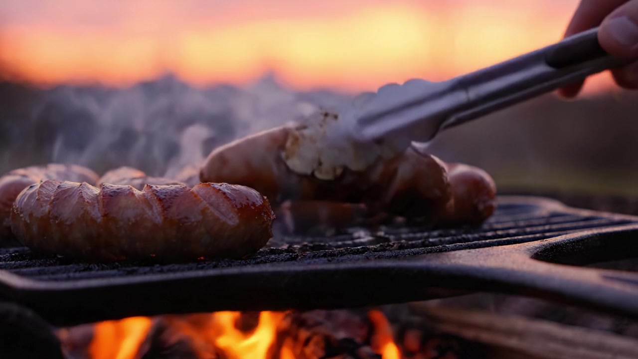Salsicce alla griglia che sfrigolano sul barbecue con uno sfondo di tramonto colorato, creando un'atmosfera accogliente e saporita per la cucina all'aperto.